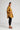 A person with long hair tied up is wearing an ATHENA NEWTON Organic Cotton Smock Blouse in ochre, along with black pants and brown shoes. They are standing sideways against a plain white background, hands in the blouse pockets. The versatile design of the smock blouse adds an effortless touch to their look.