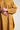 Close-up of a person wearing the ATHENA NEWTON Organic Cotton Smock Blouse in ochre, featuring long sleeves with buttoned cuffs. Their hands rest on the chest area, highlighting the versatile design and two large front pockets of this blouse against a plain light gray background.