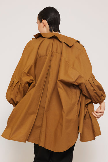 A person with slicked-back hair is wearing oversized black pants and the Organic Cotton Smock Blouse in ochre from ATHENA NEWTON. The blouse, known for its versatile design, features wide sleeves and a pleated back. Turned slightly away, the silhouette captures attention with its stylish flair.