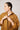 A person with wet hair styled back, wearing the Organic Cotton Smock Blouse - Ochre by ATHENA NEWTON, looks to the side while adjusting the collar. The blouse's versatile design stands out against the plain white background.