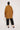 An individual wearing the ATHENA NEWTON Organic Cotton Smock Blouse in ochre, paired with black pants, stands against a plain white background, facing away from the camera.