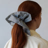 A person with long auburn hair is seen from behind, adorned with a large ATHENA NEWTON Cotton Square Scrunchie in the Railroad pattern as a hair accessory. The background is plain, and the person is dressed in a light-colored shirt.