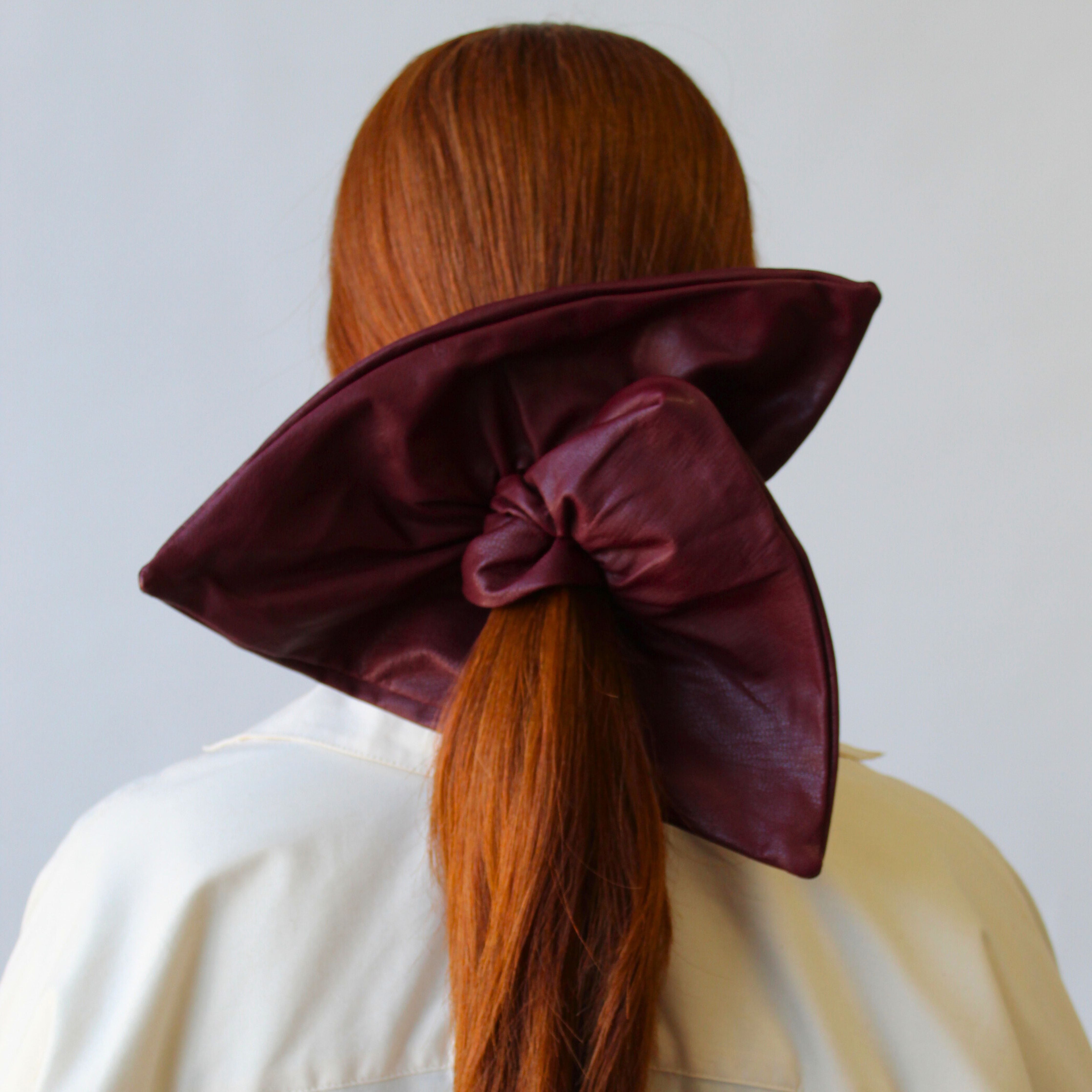 A person with long red hair is facing away, wearing the Leather Square Scrunchie in Wine by ATHENA NEWTON, tied around their neck over a cream-colored coat. The scrunchie is the focal point of the image, showcasing an elegant and stylish accessory.