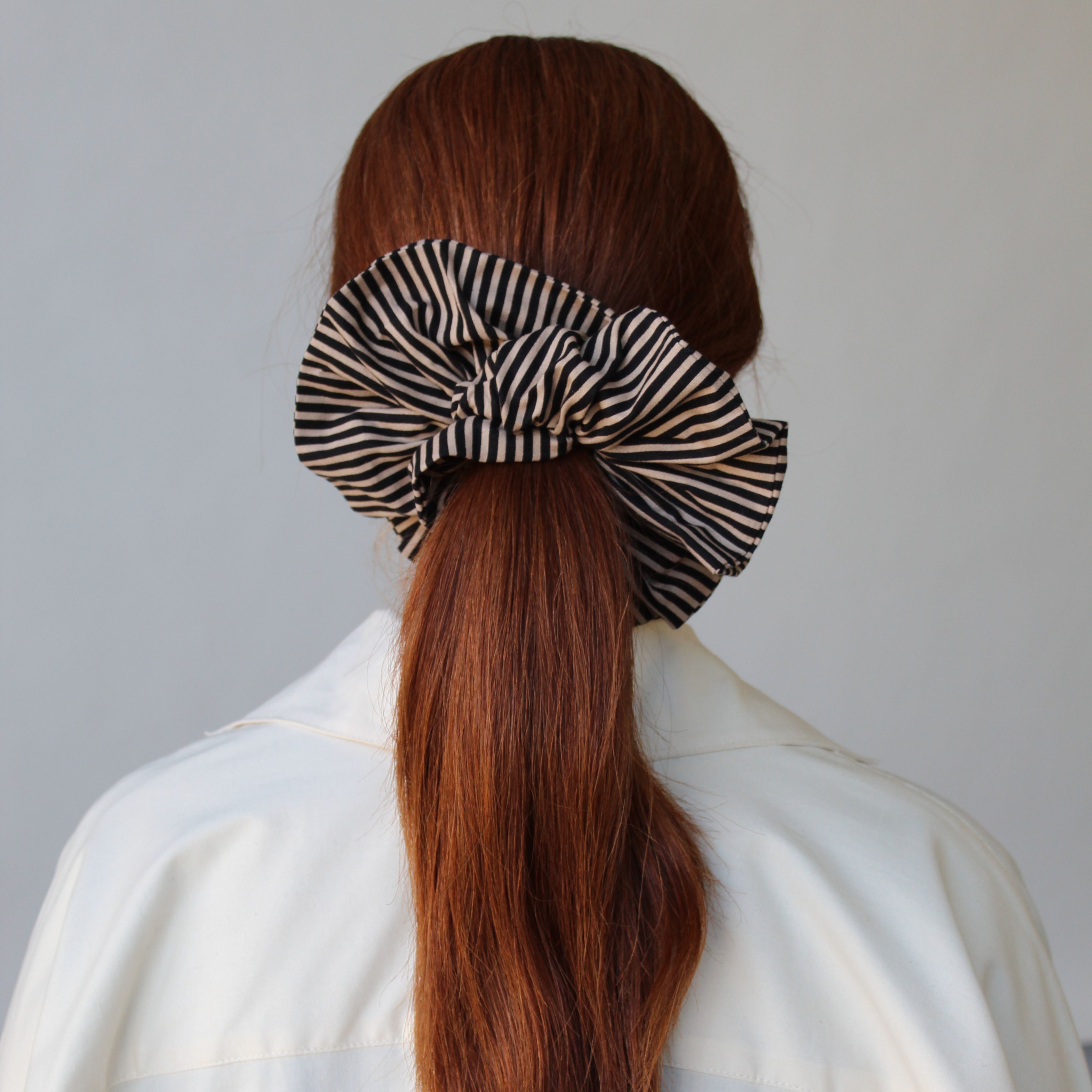 A person with long red hair is seen from behind, wearing a white shirt. Their hair is tied back with an ATHENA NEWTON Cotton Scrunchie in brown and black stripes.