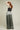 A person with long red hair stands slightly turned away, hands on their head, wearing a black sleeveless top and ATHENA NEWTON's Silk Wide Leg Pants in Sage. The oversized silhouette and relaxed fit harmonize beautifully with the plain light beige background.