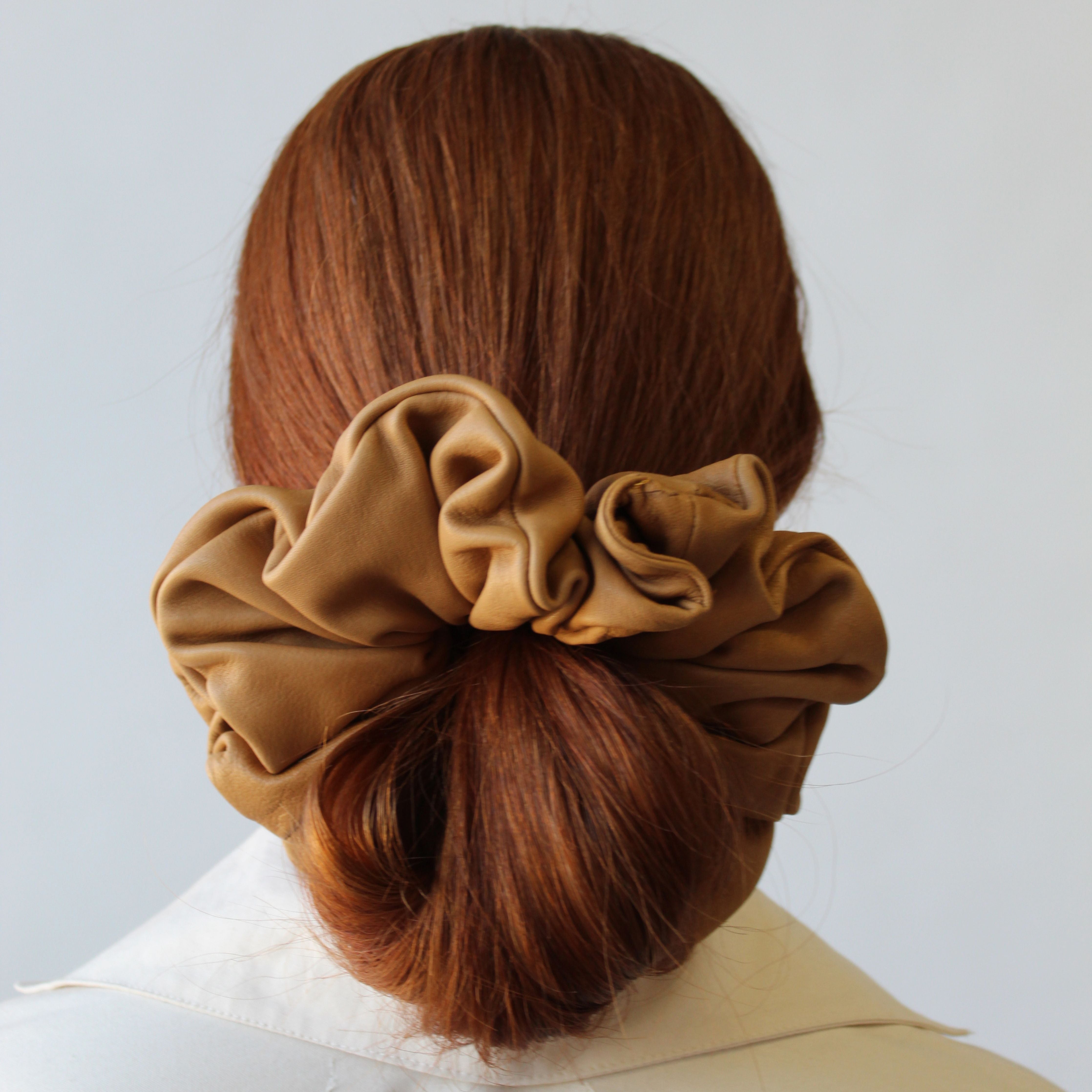 A person with long auburn hair is shown from behind, styled in a low bun secured with the ATHENA NEWTON Leather Scrunchie in Tawny. They are wearing a cream-colored collared jacket against a plain light gray background.