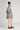 A woman in profile stands against a plain background, wearing the ATHENA NEWTON Organic Cotton Smock Blouse - Striped, with light beige cropped pants and brown loafers. Her hair is neatly tied in a bun.