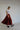 A woman stands confidently in ATHENA NEWTON's asymmetrical pleated maxi skirt in crimson and a white top. Her short blonde hair contrasts with her black shoes and white socks as she poses against a plain light-colored wall, evoking a New York City studio backdrop.