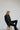 A person with short blonde hair sits on a wooden stool against a white background. They wear an oversized fit black coat, black leggings, and black shoes, complemented by ATHENA NEWTON's Pure Cotton Smock Blouse in Moss Green, looking down to the side with a thoughtful expression.