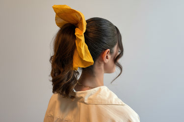 A woman with wavy brown hair styled in a ponytail is secured with the Luxurious Silk Oversized Scrunchie in Golden Mustard by ATHENA NEWTON. She wears a light top, and the softly lit background adds subtle texture.