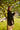 In a sunlit park beneath a tree, a person with long hair wears the ATHENA NEWTON Eyelet Smock Blouse in black. They face the camera with a backdrop featuring a fence, grass, and distant buildings.