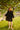 Barefoot on the grass, a person stands under a tree in ATHENA NEWTON's Eyelet Smock Blouse - Black and holds a white jacket. The sun casts warm, dappled shadows through the leaves, while a wooden railing and building subtly frame the serene scene.