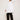 Against a plain background, someone models an oversized flowy fit, wearing ATHENA NEWTON's Cotton Smock Blouse in white with large pockets and black pants. They pair it with gold braided heeled sandals, gently touching their hair with their right hand.