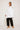 Against a plain background, someone models an oversized flowy fit, wearing ATHENA NEWTON's Cotton Smock Blouse in white with large pockets and black pants. They pair it with gold braided heeled sandals, gently touching their hair with their right hand.