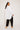 A person with long dark hair poses against a plain background, wearing an ATHENA NEWTON Cotton Smock Blouse - White and black pants. They turn slightly to the side, looking over their shoulder, with beige heeled sandals adding the final touch.