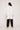 A person with long dark hair stands facing away, wearing an oversized Cotton Smock Blouse - White by ATHENA NEWTON, black pants, and beige shoes with a slight heel. The background is a plain light color.