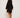 Dressed in an ATHENA NEWTON Eyelet Smock Blouse - Black with embroidered floral patterns, a person in beige sneakers stands facing away yet glances over their shoulder. The wide-sleeved blouse appears graceful against the plain, light backdrop.