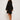 Dressed in an ATHENA NEWTON Eyelet Smock Blouse - Black with embroidered floral patterns, a person in beige sneakers stands facing away yet glances over their shoulder. The wide-sleeved blouse appears graceful against the plain, light backdrop.
