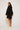Dressed in an ATHENA NEWTON Eyelet Smock Blouse - Black with embroidered floral patterns, a person in beige sneakers stands facing away yet glances over their shoulder. The wide-sleeved blouse appears graceful against the plain, light backdrop.
