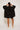 An individual with long hair tied back, facing away from the camera, adjusts their Eyelet Smock Blouse - Black by ATHENA NEWTON worn over black shorts. Beige sneakers complete the look against a plain white background.