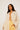 A person stands confidently wearing an ATHENA NEWTON Eyelet Smock Blouse - Ivory with no shirt underneath, paired with high-waisted light yellow pants. The floral embroidery catches the light against a plain white background.